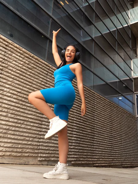 Ground level of merry young Hispanic woman in blue sportswear and white sneakers raising arm and winking while listening to music in wireless headphones and dancing outside modern building on city street