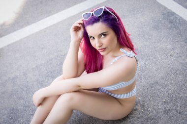 Şık güneş gözlüklü, saçlarına dokunan ve asfalt yolda oturan genç bir kadının yan görüntüsü.