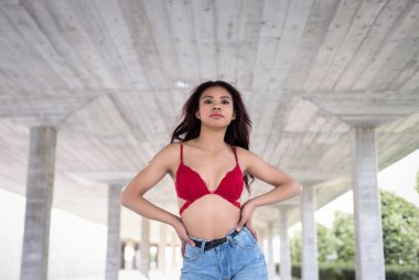 Kendine güveni düşük, modaya uygun giyinmiş genç bir kadın avluda dikilirken kameraya bakıyor.