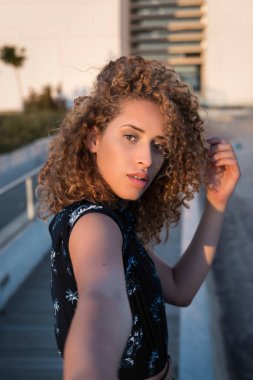 Kıvırcık saçlı güzel bir kadın güneşli bir yaz gününde inşaat ile şehrin caddesinde dikilirken kendi portresini çekiyor.