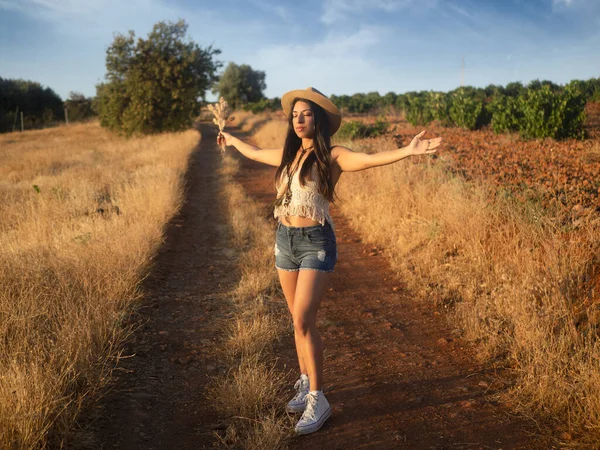 Full body young woman in casual clothes with hat spreading arms and inhaling fresh air while standing on countryside road in dry field