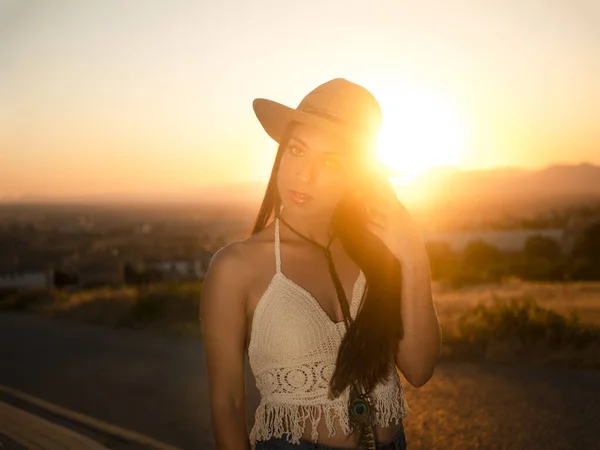 Relaxed female in crochet top and straw hat touching long dark hair and looking at camera against sunset sky in weekend evening in countryside