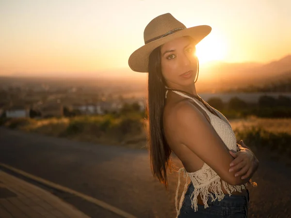 Side view of woman in casual clothes and straw hat with long dark hair crossing arms and looking at camera against bright sundown sun in countryside