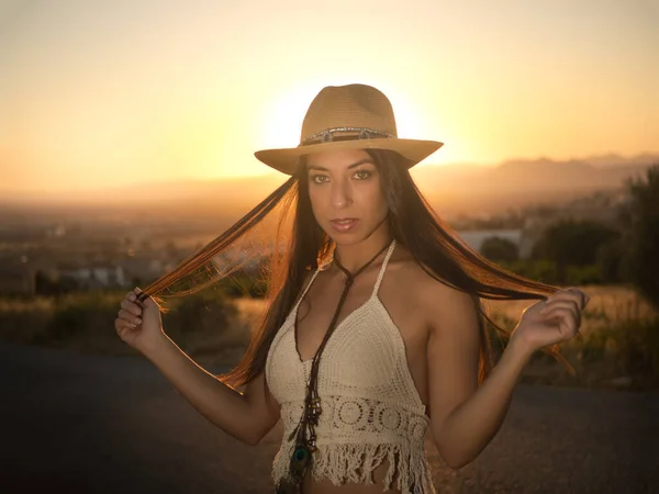 Calm woman in stylish crochet top and straw hat touching long dark hair and looking at camera on blurred background of countryside and sundown sky