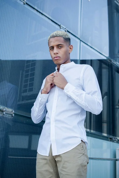 Serious African American male in classy outfit looking at camera while standing near modern glass building in city and buttoning white shirt