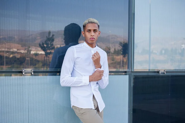 Elegant African American male in white shirt looking at camera while standing near modern glass building in city on summer day