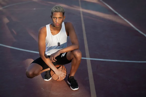Full body of serious African American male athlete in activewear looking at camera while squatting on sports ground with basketball