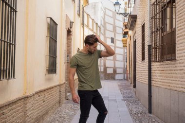 Side view of metrosexual male with muscular body in t shirt and jeans touching hair while standing on narrow street