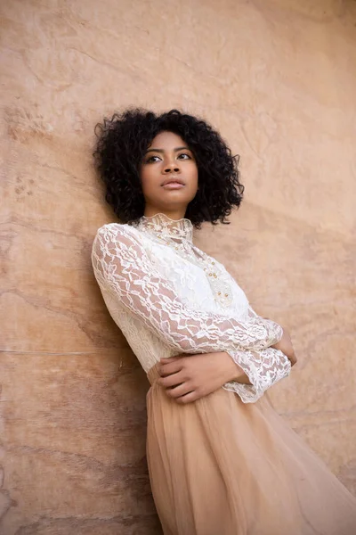 Low angle of calm young African American lady with curly dark hair in romantic white lace blouse and skirt leaning on beige wall and looking away