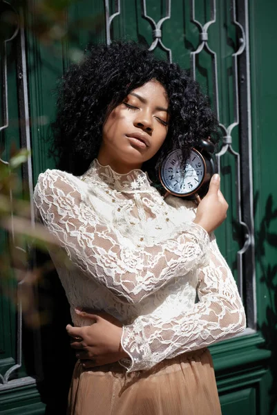 Tranquil young African American female model with curly dark hair in romantic white lace blouse standing near green wooden door with closed eyes and vintage alarm clock on shoulder