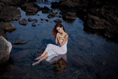 Side view of graceful young barefooted female with long dark hair in white dress sitting on stone in sea and embracing starfish in nature