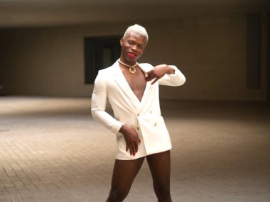 Glad androgynous African American man with short blond hair and red lips looking at camera with smile and touching elegant white jacket while standing in spacious room