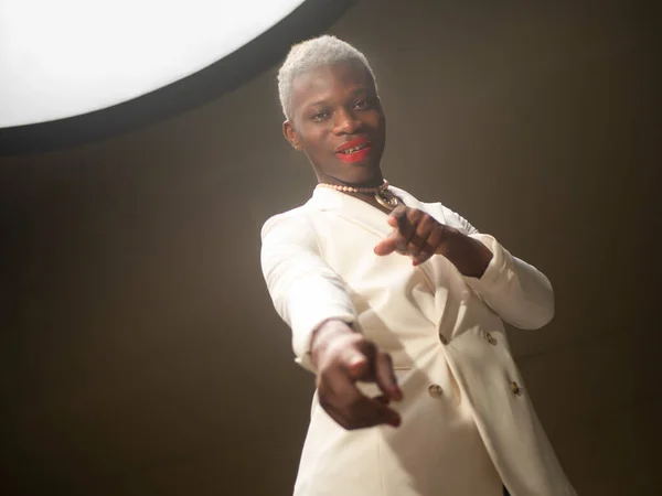 Low angle of positive African American male in elegant white jacket with short blond hair and red lips pointing at camera while standing under lamp in dim room