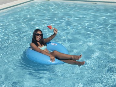 From above optimistic young Hispanic female smiling and raising arm with watermelon on stick while lying on inflatable tube in clean pool on summer day on resort