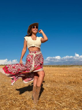 Full body optimistic woman in boho outfit looking away with smile and waving ornamental skirt while standing in dry field against cloudy blue sky on summer day