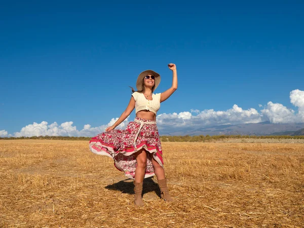 Full body optimistic woman in bohemian clothes raising arm and waving ornamental skirt while dancing in dry field against clue sky on summer weekend day
