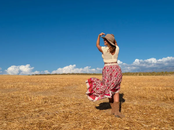Back view of merry female in boho clothes touching hat and looking away with smile while walking in dry field against cloudy blue sky on summer weekend day in nature
