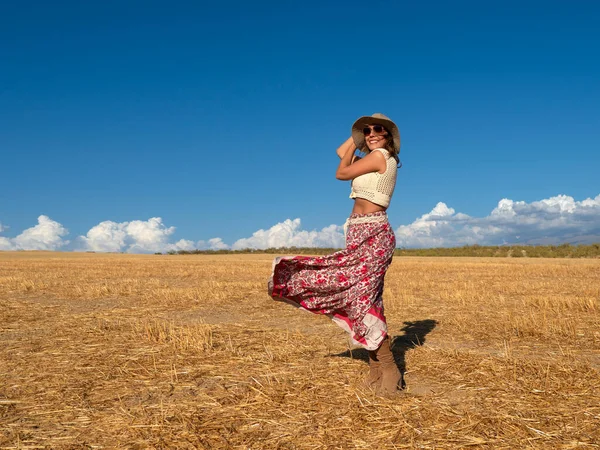 Full body cheerful woman with skirt fluttering on wind smiling and touching hat while standing on dry grass against cloudy blue sky in countryside