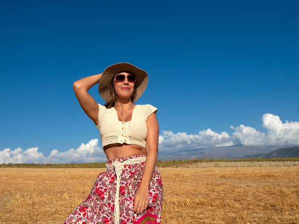 Glad female in stylish boho outfit with sunglasses touching hat and looking away with smile against cloudy blue sky on summer weekend day in dry field