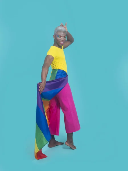 Full body happy African American man with LGBT flag wrapped around hips touching head and looking at camera with smile against turquoise background