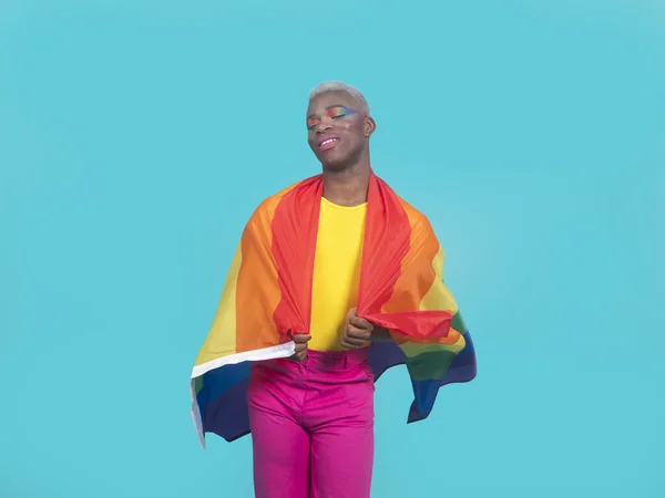 Positive African American gay with colorful makeup wrapping rainbow flag around shoulders and smiling with closed eyes against turquoise background