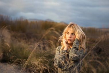 Kendine güvenen, uzun sarı saçlı ve makyajlı genç bir manken. Dokunaklı yüzler takınıyor ve gün batımında yemyeşil çayırlara bakıyor.