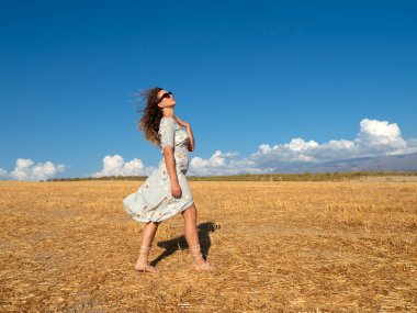 Güneş gözlüklü, yaz elbisesi takmış, saçları rüzgarda savrulan kırsal alandaki altın çimlerde güneş ışığı altında genç ve narin bir esmerin yan görüntüsü.