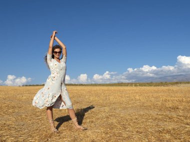 Güneş gözlüğü ve yaz elbisesi takmış genç esmerler kırsal alandaki altın çimlerde rüzgarda dikilirken kollarını kaldırıyorlar. Arka planda mavi gökyüzü.