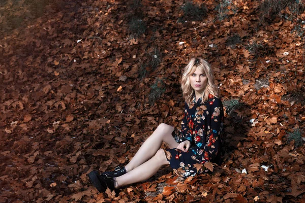 秋天的树林里 穿着黑色长裙 脚踩着靴子 长满枯叶的金发女青年高角坐在地上 — 图库照片