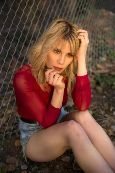 High angle of gorgeous young female model in mini denim shorts and red blouse touching long blond hair and looking away near chain link fence in countryside