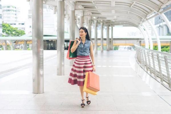 Alışveriş yaptıktan ve alışveriş poşeti taşıdıktan sonra mutlu ve gülümseyerek aldığı neşeli Asyalı genç kadın.