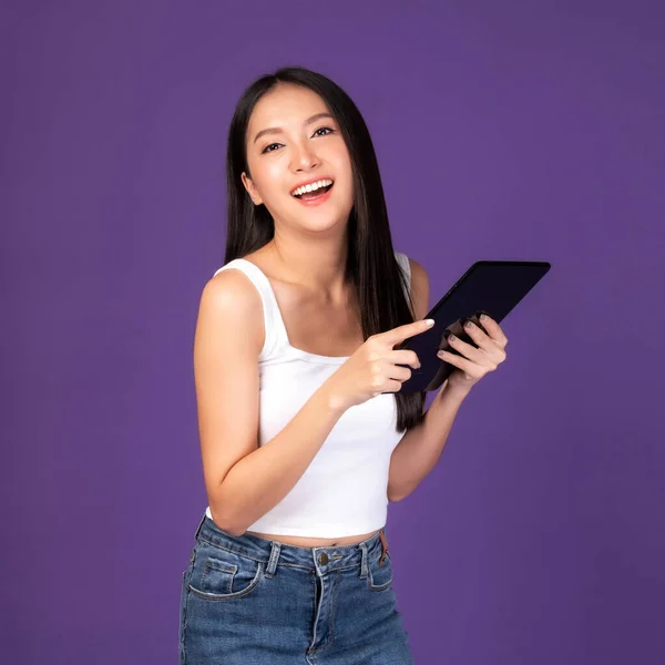 Beautiful Asian brunette woman cute girl in white tank top playing a smart phone , tablet computer isolated on purple background - mobile phone uses a online banking for pay online shopping