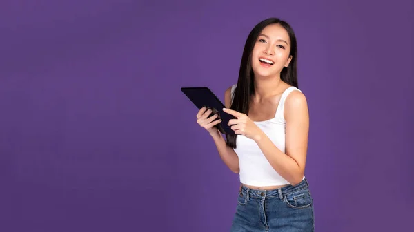 Beautiful Asian brunette woman cute girl in white tank top playing a smart phone , tablet computer isolated on purple background - mobile phone uses a online banking for pay online shopping