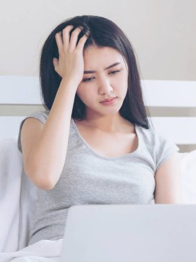 Yatakta oturan genç ve güzel bir kadın dizüstü bilgisayar kullanıyor. Ciddi ve baş ağrısı çekiyor.