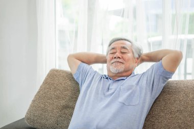 Emekli bir Asyalı son sınıf öğrencisi, gülümseyen yaşlı bir adam, evinin oturma odasındaki kanepede dinlenen yaşlı bir adam - son sınıf mutluluk konsepti.