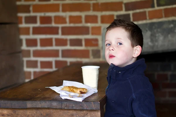 Little boy eating sausage roll and a cup of coffee — Stock Photo, Image