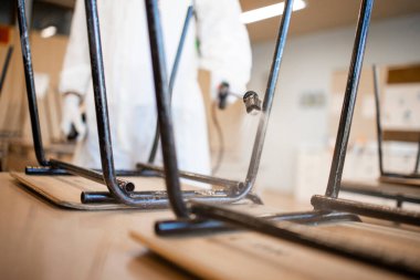 Corona virüsü salgını sırasında sınıftaki masaları ve sandalyeleri dezenfekte ve dezenfekte eden beyaz steril giysili bir adamın yakın görüntüsü.