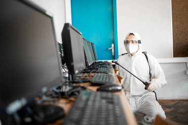 Beyaz steril koruyucu giysili bir adam Corona virüsü salgını sırasında okuldaki bilişim sınıfının klavye ve ekipmanlarını dezenfekte ediyor ve dezenfekte ediyor..