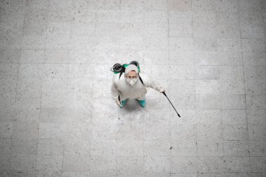 Tanınmayan beyaz kimya korumalı bir kişinin, yüksek derecede bulaşıcı korona virüsü salgılamasını engellemek için kamu alanlarını dezenfekte edişinin üst görüntüsü..