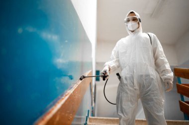 Beyaz steril koruyucu giysili bir adam Corona virüsü salgını sırasında okul sınıfındaki merdivenleri ve koridorları dezenfekte ediyor ve dezenfekte ediyor..