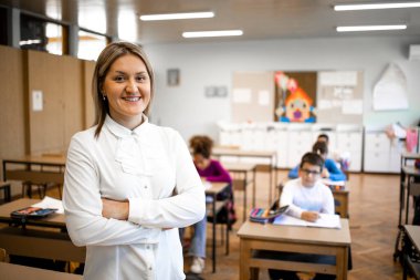 Sınıfta duran öğretmen portresi ve arka planda oturan çocuklar..