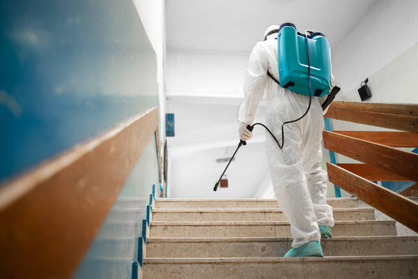 Man in white sterile protection suit disinfecting and sanitizing steps and corridors in school classroom during corona virus pandemic.