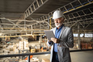 Fabrikanın CEO 'su ya da yöneticisi takım elbiseli ve kasketli olarak üretim salonunda duruyor ve şirketinin sonuçlarını kontrol ediyor. Arka planda çalışan endüstriyel fabrika içi ve forkliftler.