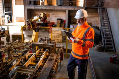 Endüstri çalışanı üretim makinelerinin yanında duruyor ve tablet bilgisayardaki üretim sonuçlarını kontrol ediyor.