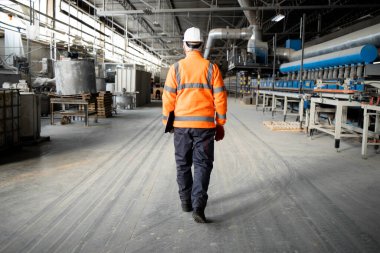 Professional factory worker or engineer in high visibility jacket and hardhat walking through industrial production hall with machinery. Factory interior. clipart