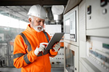 Yüksek görüş menzilli ceketli deneyimli bir fabrika işçisi ya da mühendis tablet bilgisayar kullanarak endüstriyel üretim salonundaki otomatikleştirilmiş makinenin yanında duruyor. Fabrikanın içi..