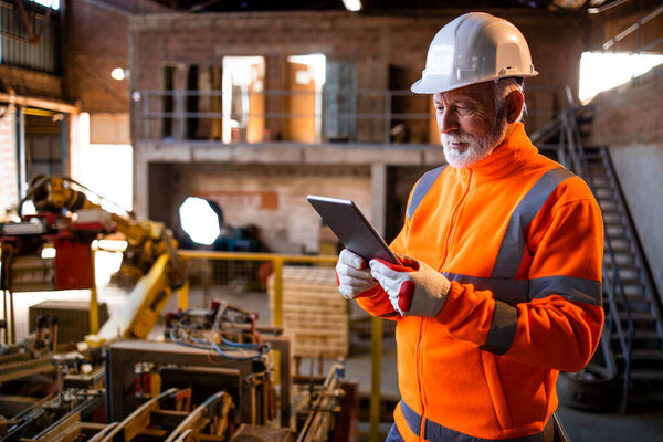 Factory worker in safety uniform and white hard hat standing inside manufacturing factory. Industrial engineer with computer standing by automated machine robotic arm and checking production process.