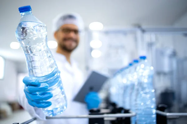 stock image Industrial production of bottled water. Technologist standing by automated filling machine and holding bottle of water.