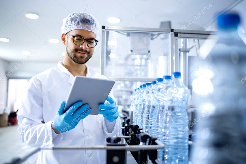 Bottling factory worker or technologist in sterile clothing standing by automated conveyor belt machine and checking water production.