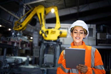 Sanayi kadın mühendisi imalat fabrikasında üretim sürecini kontrol ederken robotik kol makinesi arka planda çalışıyor..